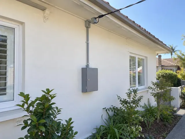 Exterior house wall with overhead service cable and point-of-attachment bracket above a grey electrical box in Manly